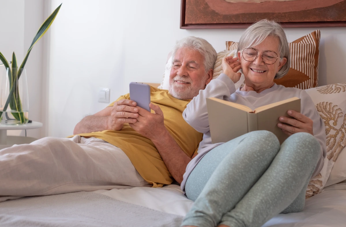 Happy Senior Couple Relaxing in Bed with a Book and Smartphone